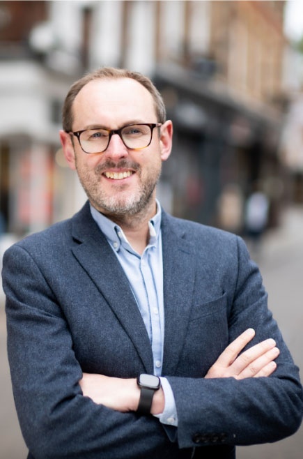 Mark Nunny stands with his arms folded and smiles into the cameqra...