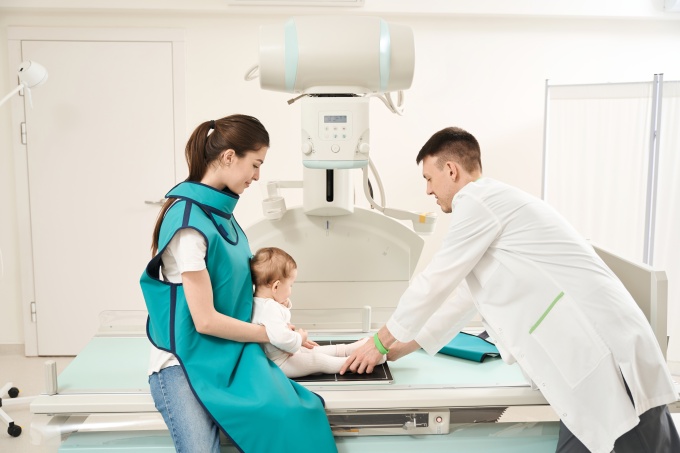 A female caregiver wearing a protective lead apron holds a baby on an X-ray...
