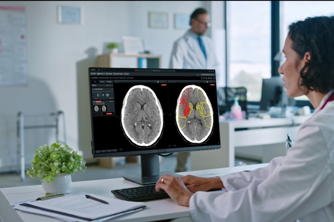 Image: A doctor sits in front of a computer monitor displaying two brain CT...