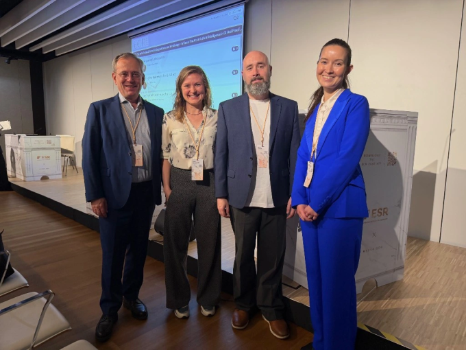 Group photo of four speakers standing in front of a stage with a presentation...